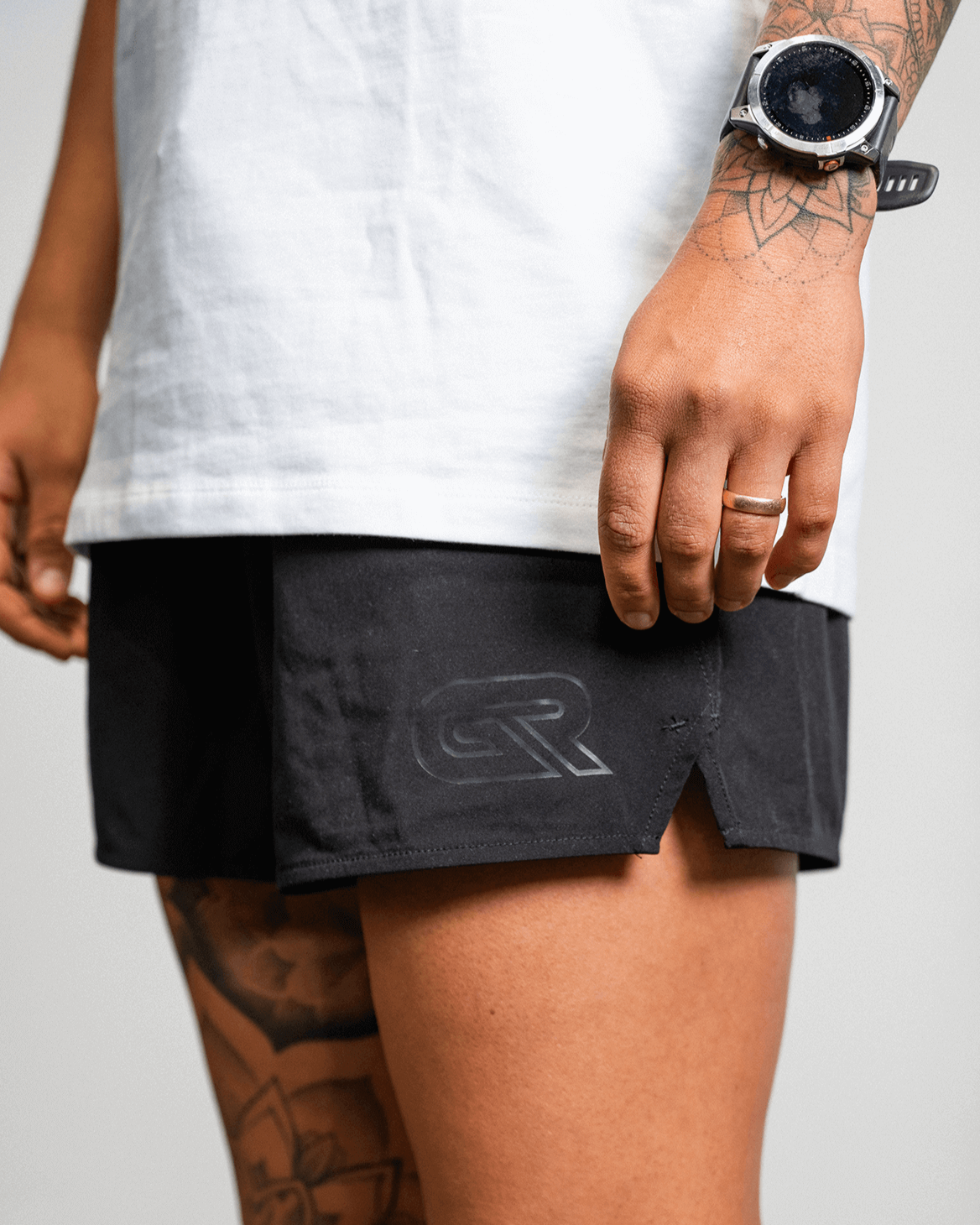 Person wearing black athletic shorts with a logo, white shirt, and tattoos on arms and legs.